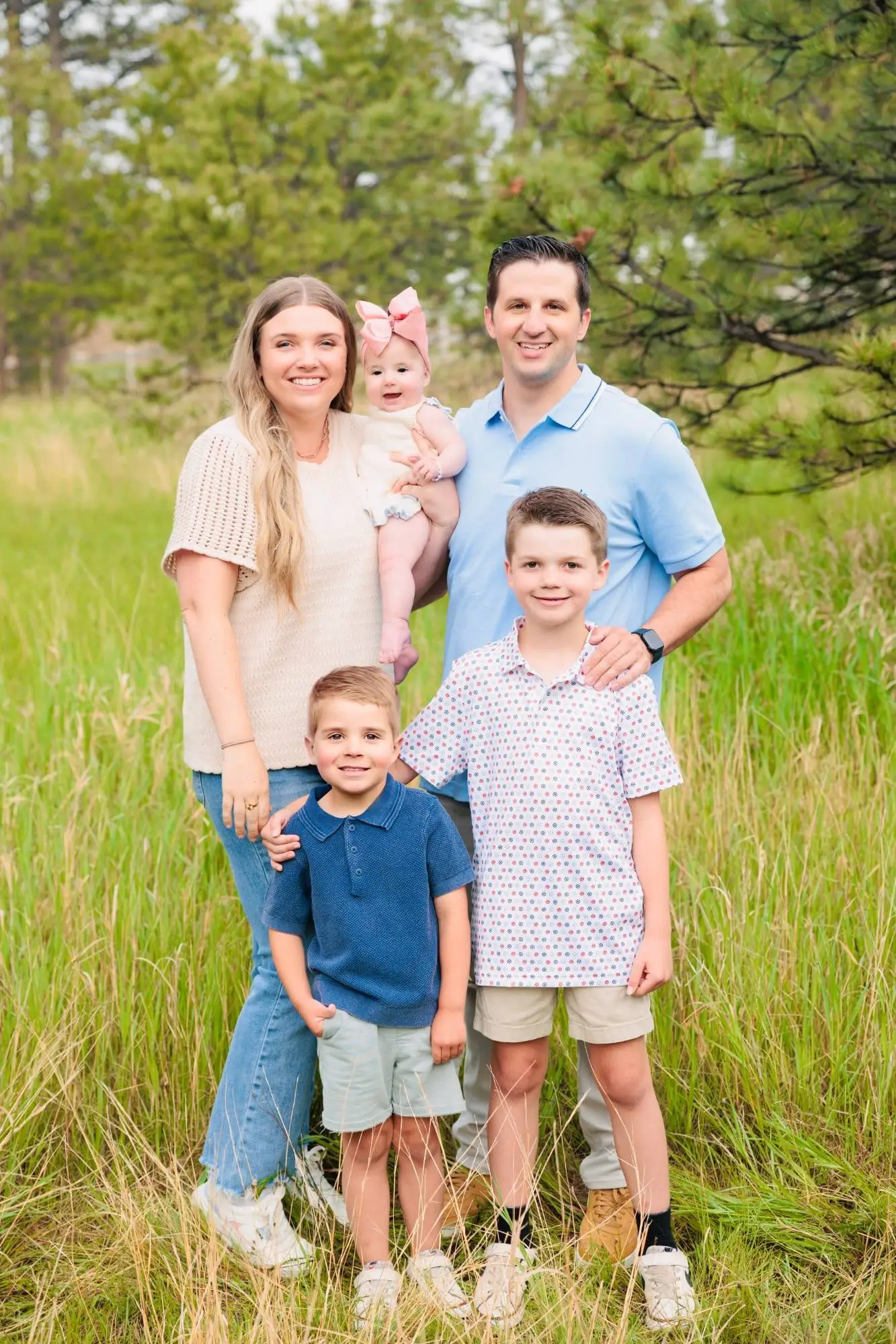 Our family of five smiling in a grassy meadow among pine trees, Mom and Dad holding our baby girl with the two boys in front