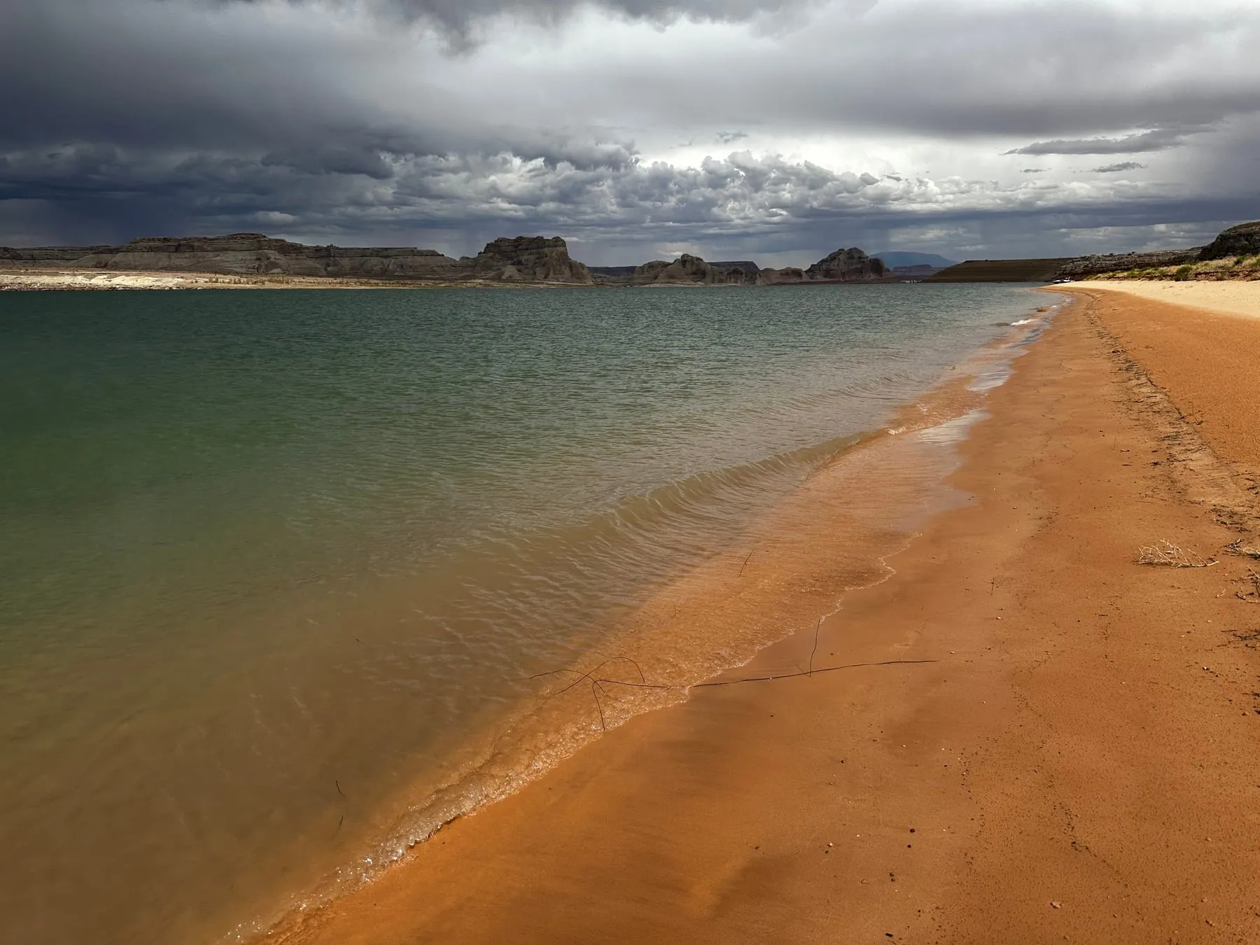 Clear green lake water lapping a red-sand beach under stormy clouds in Utah