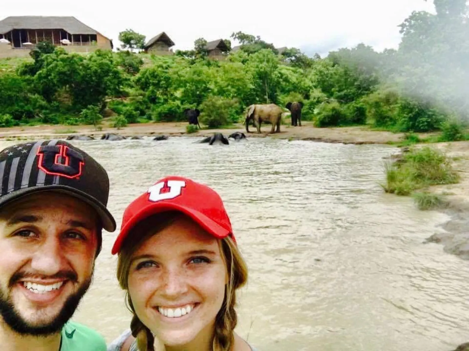 Michael and Chantelle taking a selfie by a river at Mole National Park, with elephants bathing behind them